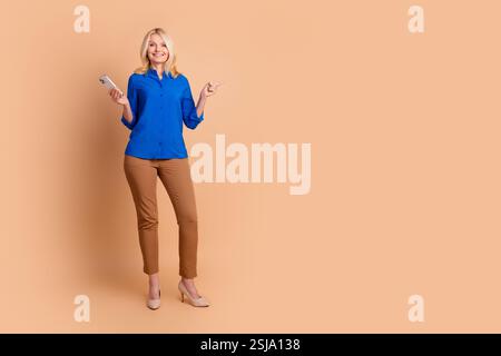 Elegant Mature Woman in Blue Shirt Using a Smartphone and Gesturing Upwards Against a Beige Background Stock Photo