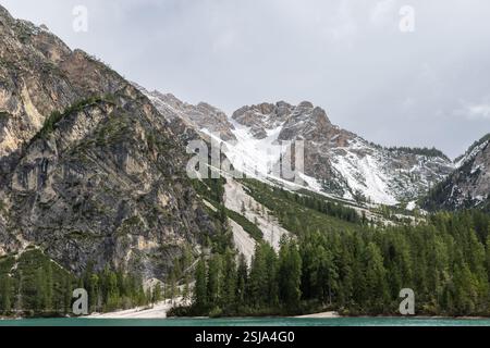 Steep rocky cliffs and snow-covered ridges rise above the lush evergreen forests and turquoise shoreline of Lake Braies, creating a dramatic alpine la Stock Photo
