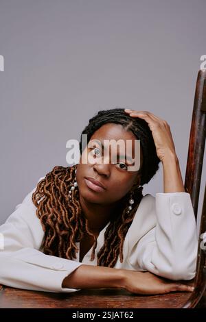 Portrait of African American woman leaning on wooden chair, wearing white shirt with thoughtful expression, dreadlocks resting on shoulders, head supported by her hand Stock Photo