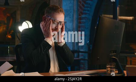 Caucasian businessman guy business man male worker working computer at night overtime office late time deadline feel headache pain head ache migraine Stock Photo