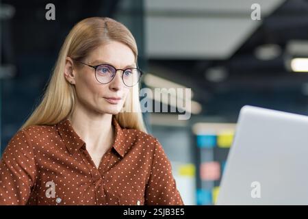 Mature woman close-up at workplace working with laptop, businesswoman looking at computer screen while sitting at desk, confident and concentrated. Stock Photo