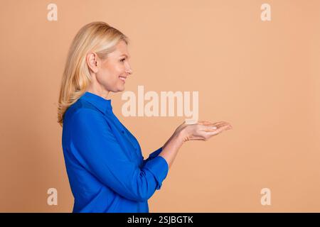 Smiling mature woman wearing a stylish blue shirt gesturing with both hands against a beige background, exuding elegance and charm. Stock Photo