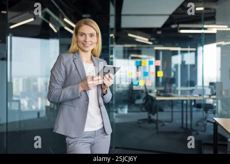 Portrait of a successful mature businesswoman with a tablet computer in her hands. Office worker in a business suit smiling and looking at the camera, standing near the window inside the office. Stock Photo