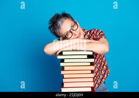 Charming Woman With Glasses Leaning on a Stack of Books Against a Bright Blue Background, Expressing Leisure and Lifestyle Stock Photo