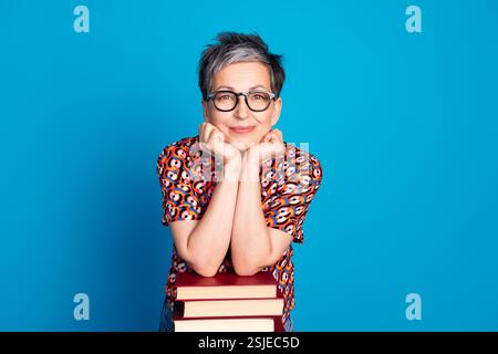 Smiling Mature Woman with Glasses in Printed Shirt Leaning on a Stack of Books Against a Blue Background Stock Photo