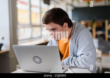 Concentrated young man with Down syndrome sitting and studying indoors at school, using laptop Stock Photo