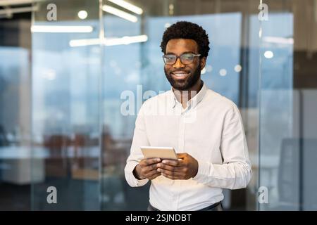 Portrait of a young successful businessman inside the office at his workplace. The man is smiling and looking at the camera, holding a tablet computer in his hands. Stock Photo