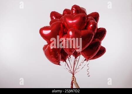 A hand holding a large bunch of red heart-shaped balloons against a clean white background, symbolizing love, romance, and celebration. Stock Photo
