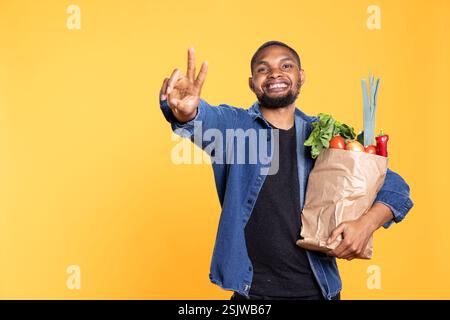 African american cheerful guy showing peace symbol in studio, feeling excited while he carries bag full of homegrown groceries. Young adult does a peaceful gesture against yellow background. Stock Photo