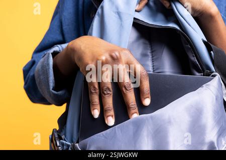 BIPOC woman putting laptop in backpack, preparing to start job shift, isolated over studio background. African american employee packing notebook, going to work, close up shot Stock Photo