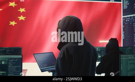 Chinese hacker working in governmental hacking room with Chinese flag on a big screen. Having the mission on inflicting fear and misinformation among the population. Dictatorship propaganda. Camera B. Stock Photo