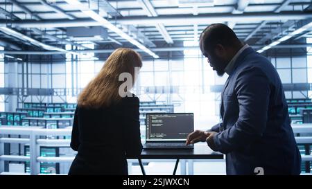 Team of developers doing maintenance checkup using laptop to prolong data center hardware life span. IT support staff members in server hub monitor server infrastructure using notebook, camera B Stock Photo