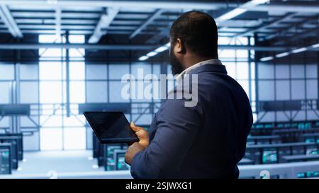 IT operator in server room workspace using tablet to patch network issues affecting transfer rates. Engineer in data center fixing errors affecting server rows rigs, camera A Stock Photo