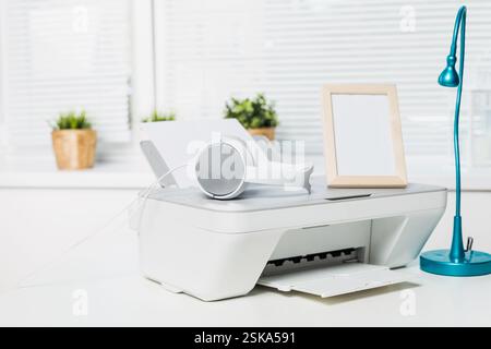 Home office setup featuring a printer, headphones, lamp, and empty frame on a desk with natural light Stock Photo