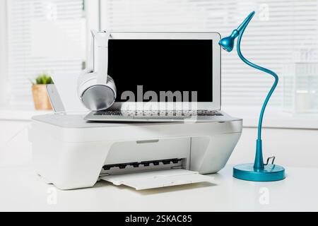 Modern workspace setup with laptop, printer, headphones, and desk lamp in bright office environment Stock Photo