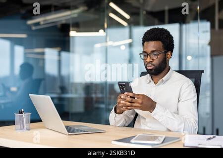Serious focused man using app on phone. Businessman thinking sitting at desk in middle of office, typing text message on smartphone, and browsing online internet pages. Stock Photo
