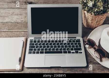 Workspace setup with laptop, notebook, sunglasses, and hat on wooden table Stock Photo