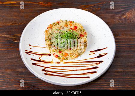 A vibrant rice dish featuring mixed vegetables is elegantly plated on a white dish. Drizzled with a savory sauce, the meal is set against a textured w Stock Photo