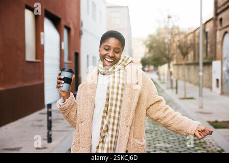 Carefree young woman laughing and holding a reusable coffee cup while walking in the street Stock Photo