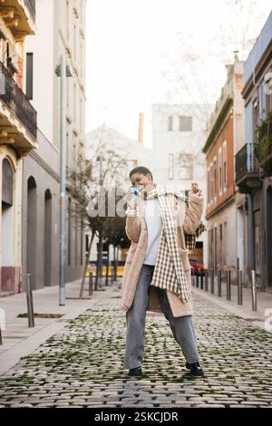 Fashionable young woman sips her coffee and dances down a charming cobblestone street Stock Photo
