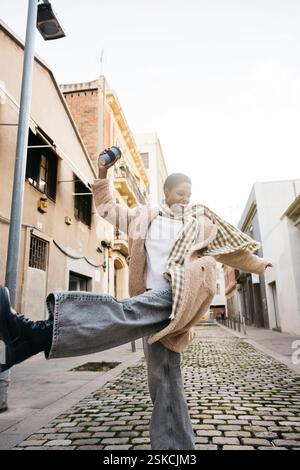 Carefree young woman dancing in the street while holding a reusable coffee cup, enjoying a sunny day in the city Stock Photo