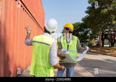 Engineers discussing logistics plans at a shipping yard, analyzing blueprints and container arrangements. Stock Photo