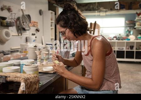 Close-up of a woman sitting at a table in a pottery studio glazing making a pot Stock Photo