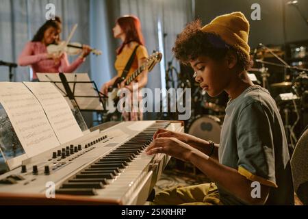 Side view of African American boy concentrated on laying keyboard while his friends chatting in blurred background Stock Photo