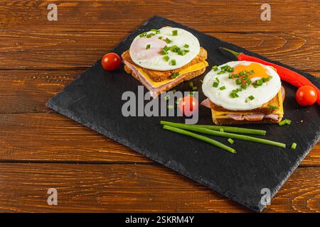 Two tasty breakfast sandwiches are served on a black slate plate. Each sandwich features crispy bread, ham, and a perfectly fried egg, garnished with Stock Photo
