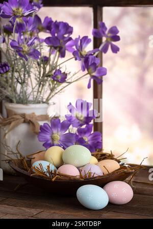Easter eggs in a bowl with purple daisies on a wooden table next to window with colorful springtime background Stock Photo