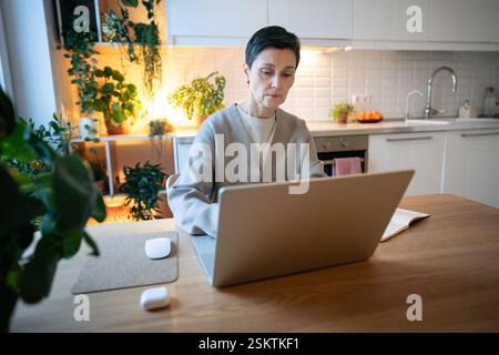 Thoughtful busy exhausted woman working on project at laptop in kitchen at home, facing deadlines Stock Photo