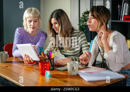 Three young creative women collaborating in a modern office, discussing ideas using a digital tablet and taking notes. Teamwork and innovation in a pr Stock Photo