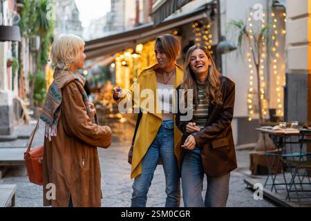 Three fashionable young women laughing and engaging in conversation while walking on a beautifully lit city street in the evening. Warm lights create Stock Photo