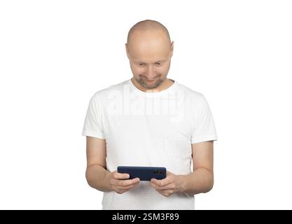 A bald Caucasian man with a short beard is wearing a white t-shirt and looking down at his smartphone with a slight smile. Stock Photo