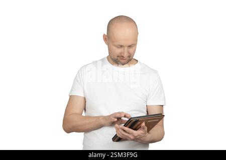 A bald Caucasian man with a short beard is wearing a white t-shirt and interacting with a tablet that has a brown cover. Stock Photo