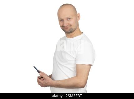 A bald Caucasian man with a short beard is standing in a relaxed pose, holding a smartphone and smiling confidently. Stock Photo