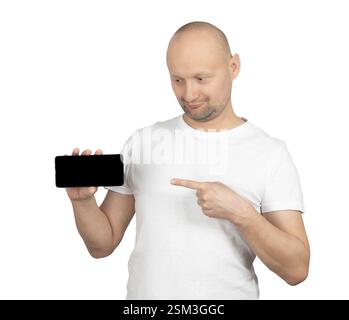 A bald Caucasian man with a short beard is wearing a white t-shirt and holding a smartphone in one hand while pointing at it with the other. Stock Photo