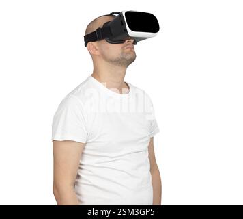 A bald Caucasian man in a white t-shirt stands against a neutral background. He is wearing a VR headset and looking upwards, appearing fully engaged Stock Photo