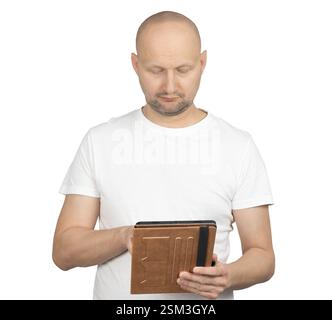 A middle-aged bald man wearing a white t-shirt is using a tablet with a brown cover. He is looking down at the screen with a focused expression. Stock Photo