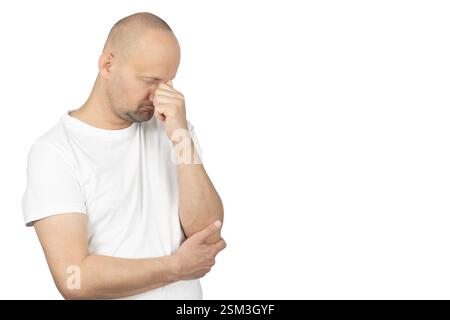 A bald Caucasian man in a white t-shirt stands against a neutral background. He rubs his forehead with his fingers, appearing deep in thought Stock Photo
