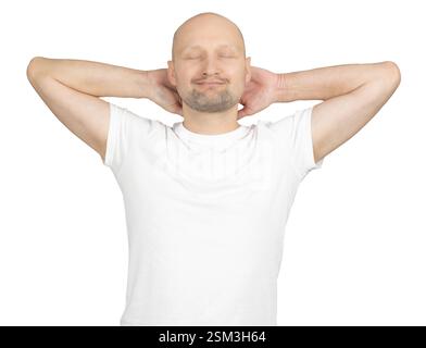 A Caucasian bald man wearing a white t-shirt is standing against a plain background. He has his hands placed behind his head and eyes closed Stock Photo