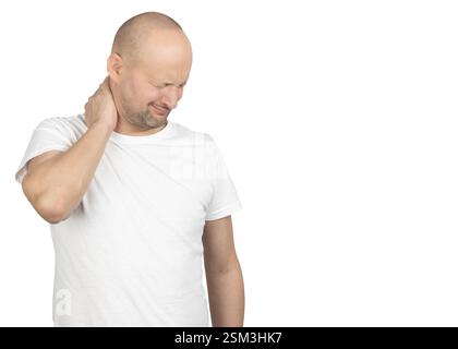 A bald Caucasian man in a white t-shirt stands against a neutral background. He rubs the back of his neck with one hand Stock Photo