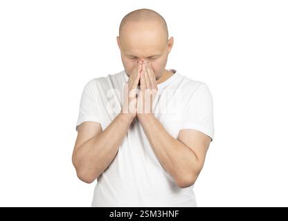 A bald Caucasian man in a white t-shirt stands against a plain background. He covers his face with both hands, expressing deep concern, stress Stock Photo