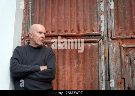 handsome bald man leaning against a rusty door with arms folded Stock Photo