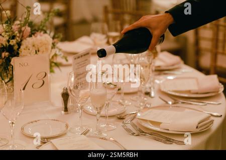 A waiter pours champagne into glasses at an elegantly set table during a formal gathering in a decorated venue. Stock Photo