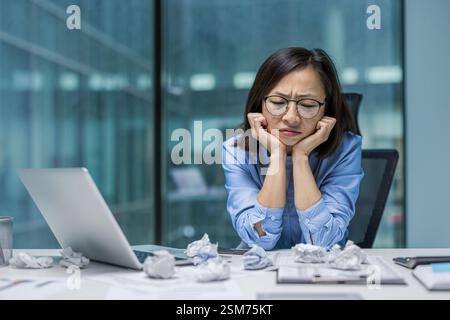 Disappointed and sad Asian woman sitting inside the office near documents and torn pieces of paper from a failed report. Woman is unhappy and nervous, working with a laptop. Stock Photo