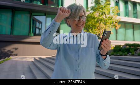 Senior happy businesswoman Caucasian female hold mobile phone gadget in city outdoors excited amazed middle-aged adult 50s old business woman win Stock Photo
