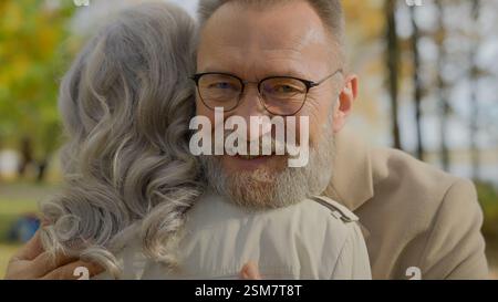 Mature couple hugging in autumn park senior gray-haired 60s aged man embracing woman happy married family embrace hug love support care romance at old Stock Photo