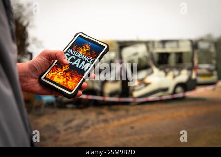 Insurance fraud investigator holding smartphone with insurance scam message with burnt vehicle in background Stock Photo