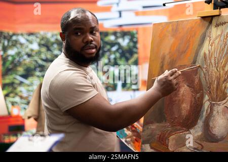 Woman master painter teaching her apprentice to use new painting techniques in art studio, sharing her skill and ideas in creating a rustic artwork on canvas. Artist helping man with paintwork. Stock Photo